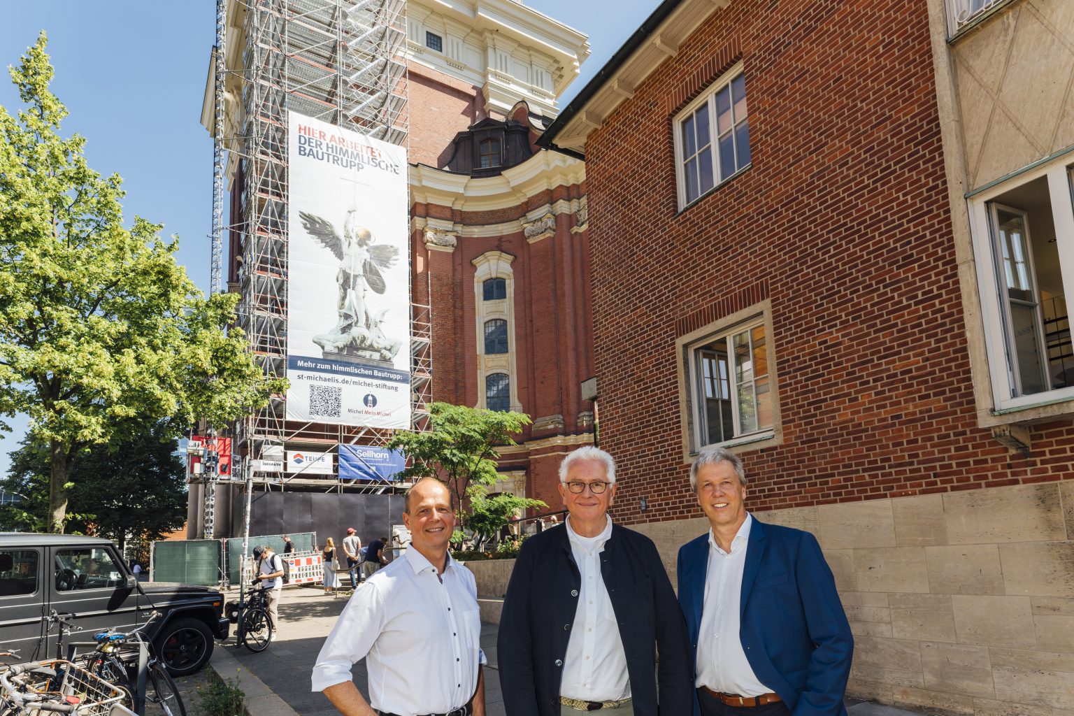 Bauleiter Uwe Pfeifer steht mit Hauptpastor Alexander Röder und Michael Kutz vor dem Banner des Michel-Turms.