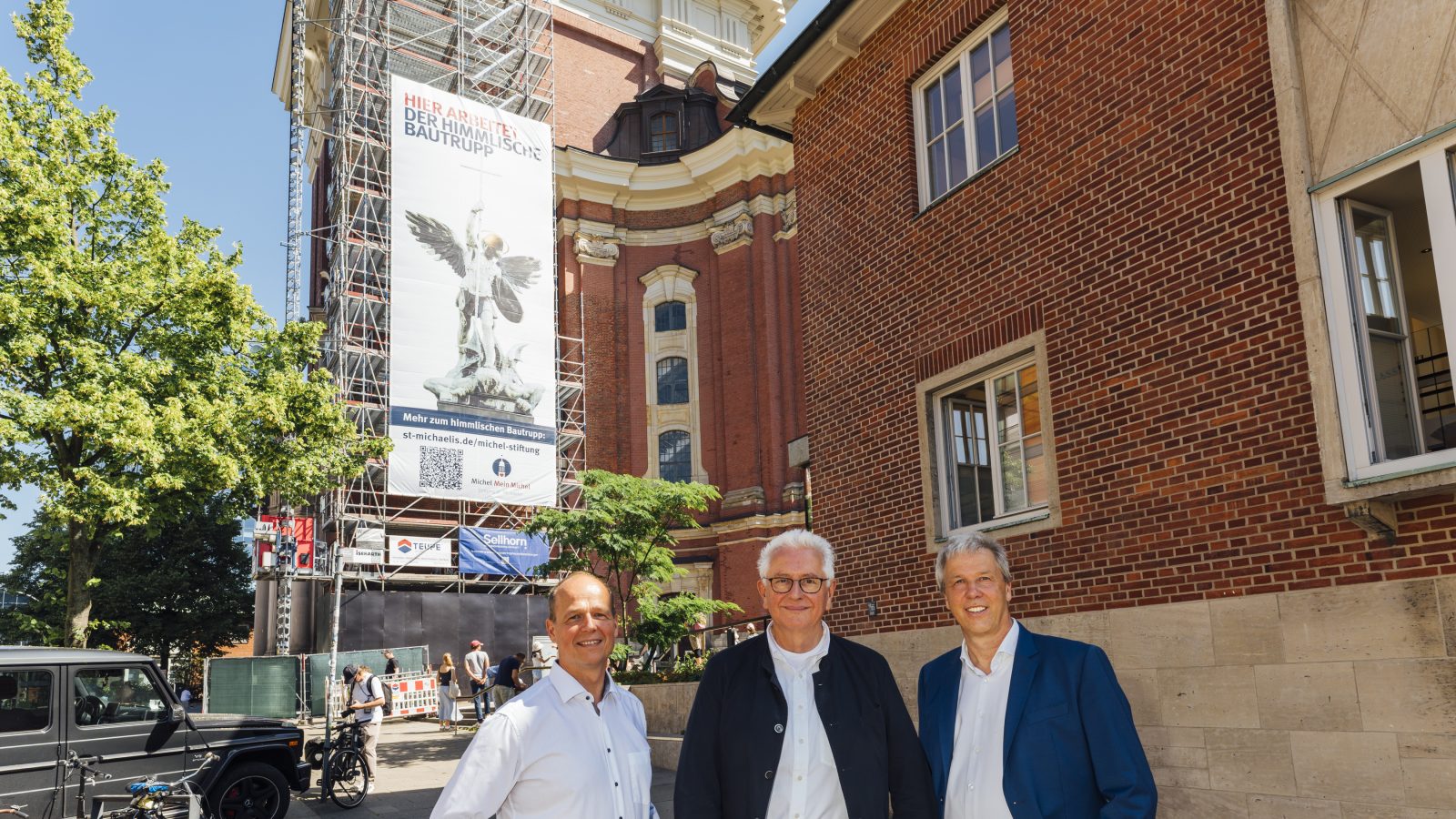 Bauleiter Uwe Pfeifer steht mit Hauptpastor Alexander Röder und Michael Kutz vor dem Banner des Michel-Turms.