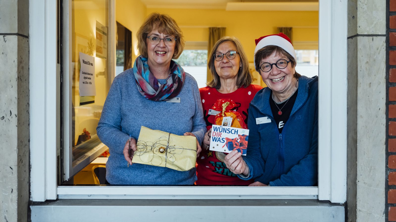Drei Frauen stehen mit einem Weihnachtsgeschenk in der Hand im geöffneten Fenster. Eine trägt eine Weihnachtsmannmütze und hält eine Postkarte der Aktion "Wünsch dir was" in der Hand